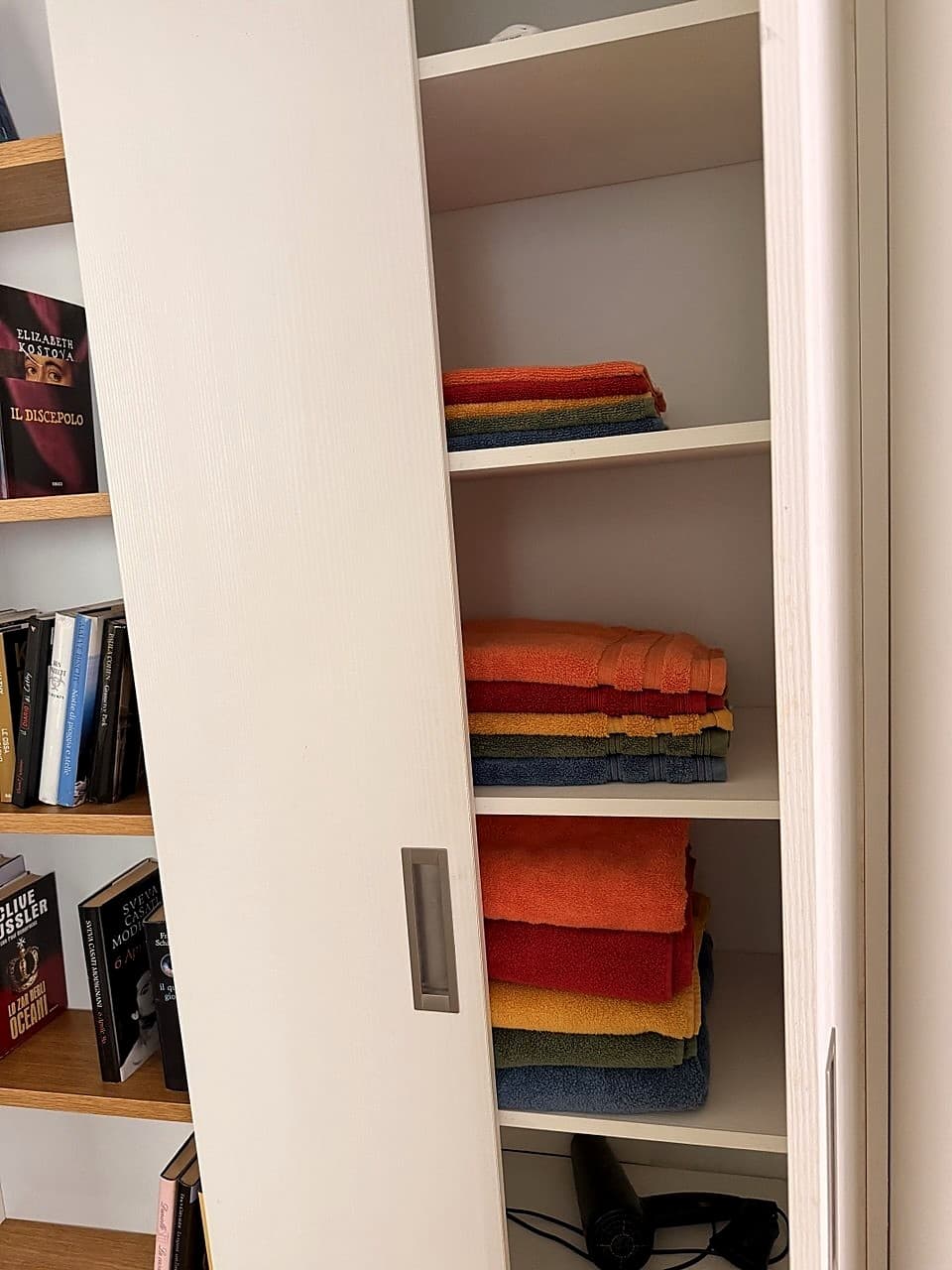 L'armadio bianco accanto al bagno, aperto · pile di asciugamani in colori vivaci (arancione, rosso, giallo, blu) impilati su tre ripiani · in basso si intravede il fon