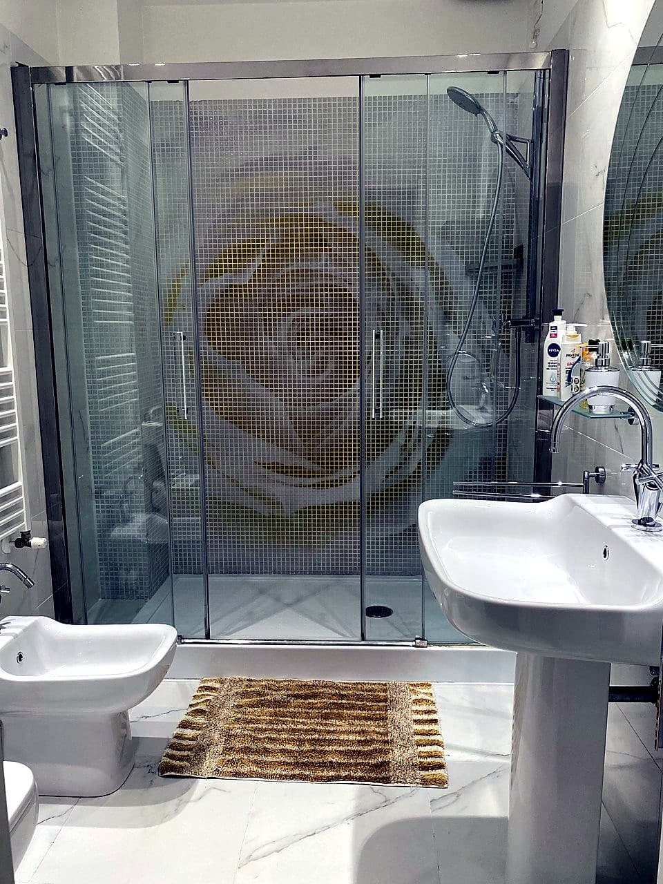 Vista d'insieme del bagno dalla zona del WC · doccia in mosaico artigianale color oro (effetto rosa stilizzata sui mosaici), bidè in primo piano, lavandino bianco sospeso a destra, pavimento e pareti in marmo bianco