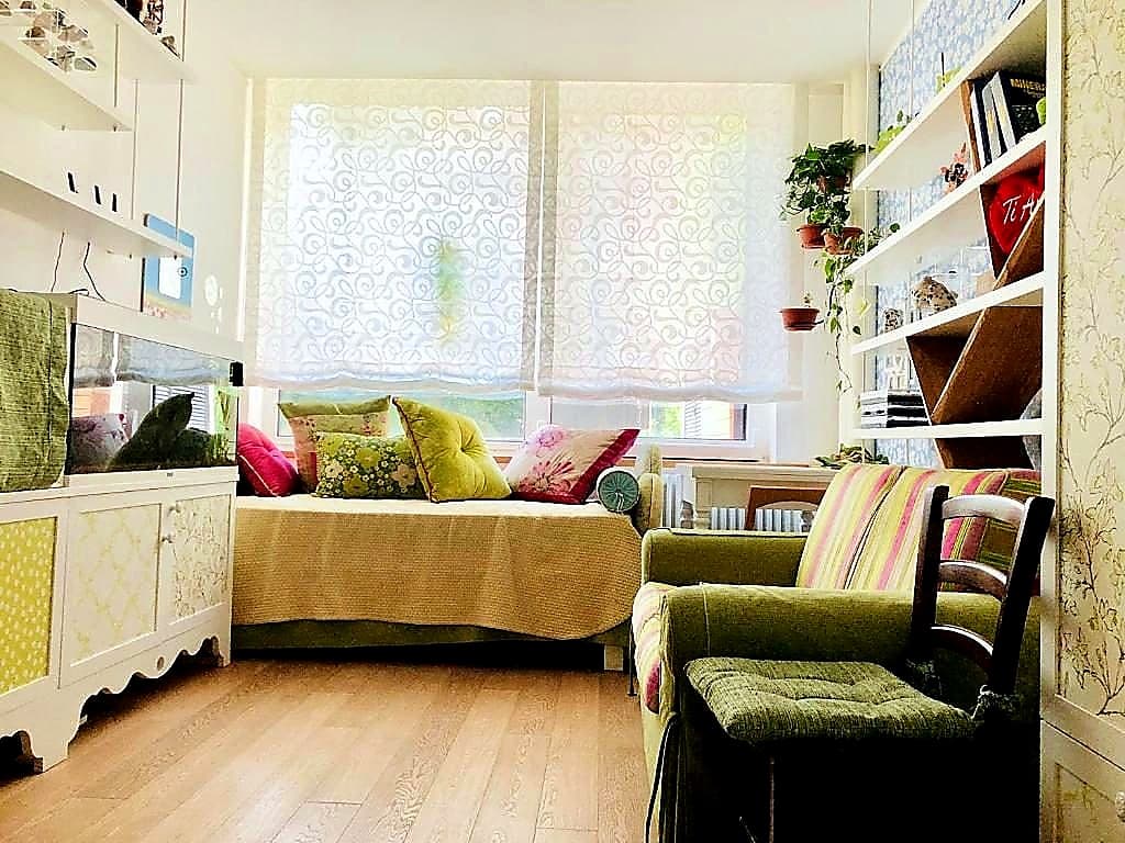 La seconda camera della Tana · divano con cuscini colorati, acquascape a sinistra, libreria a destra, scrittoio davanti alla finestra con tenda decorata