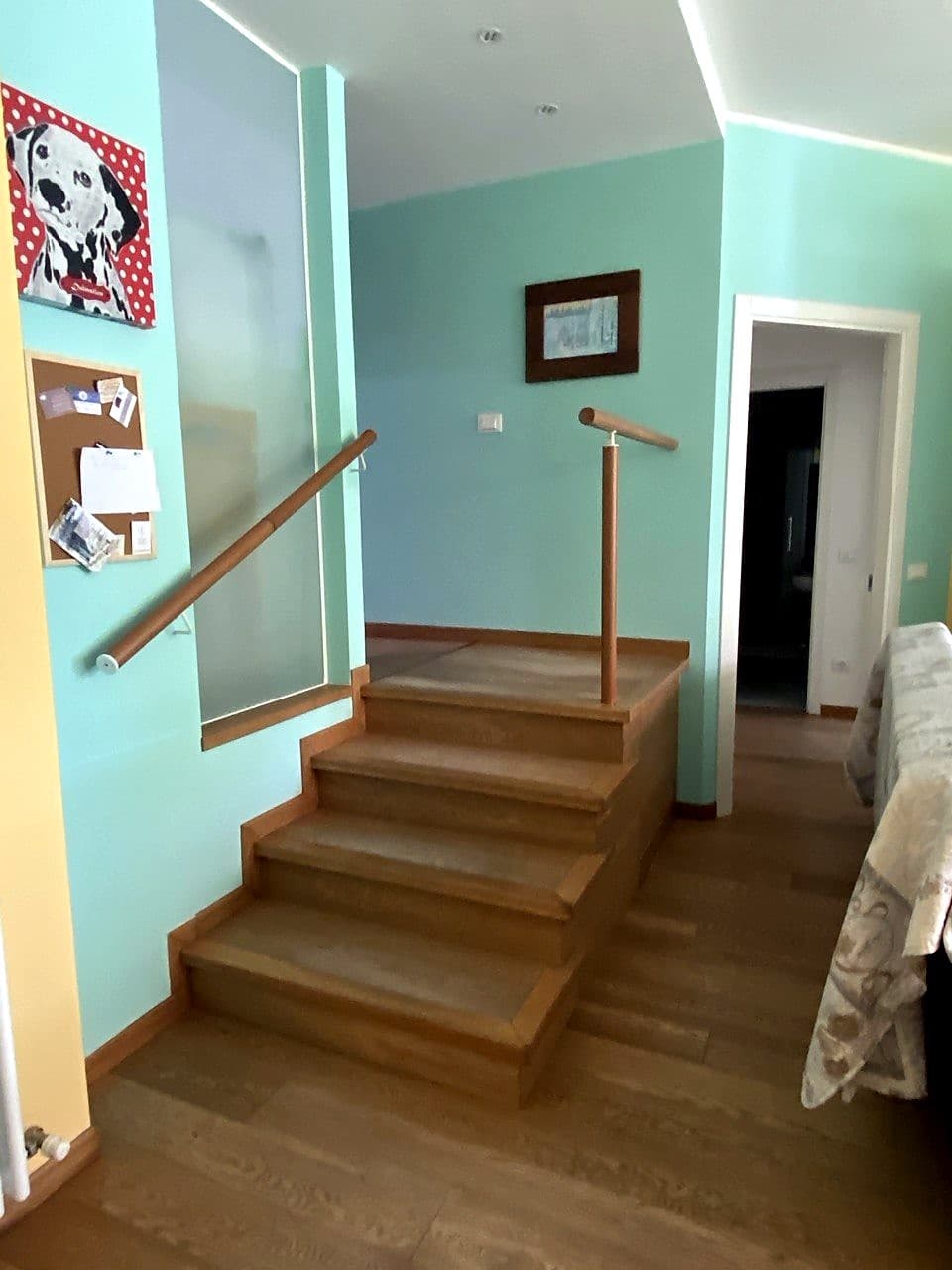 La scaletta interna di legno che separa l'ingresso e la lavanderia dal piano principale dell'appartamento · doppio corrimano (uno a parete, uno a colonnina), pareti color salvia chiaro, un dipinto pop con dalmata a pois rossi a sinistra
