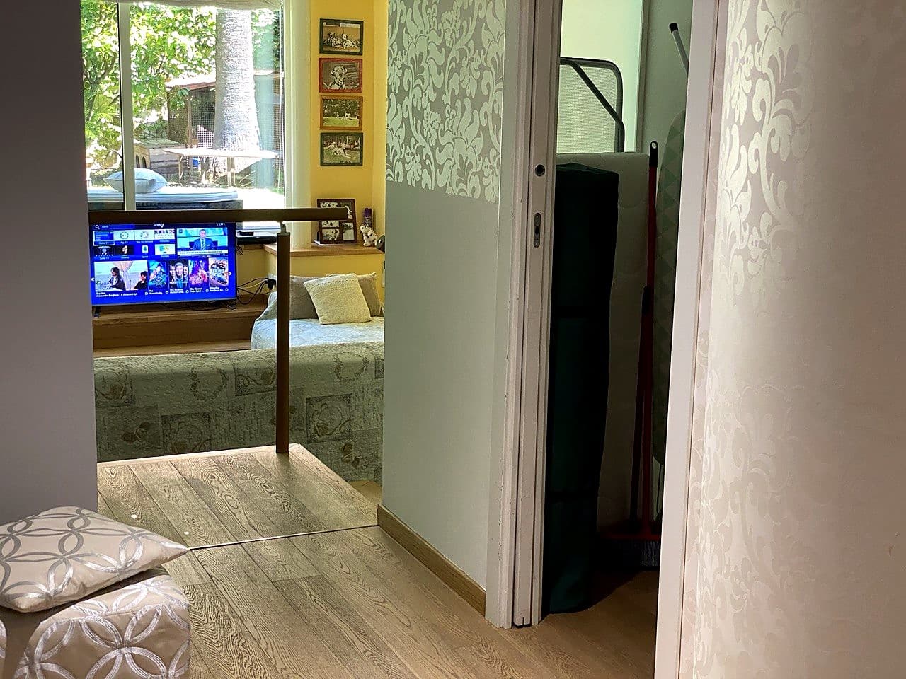 Vista dall'ingresso verso l'appartamento · la TV Smart accesa, un divano-letto preparato sotto la finestra, e sulla destra il piccolo ripostiglio della lavanderia con materassino da viaggio e scopa