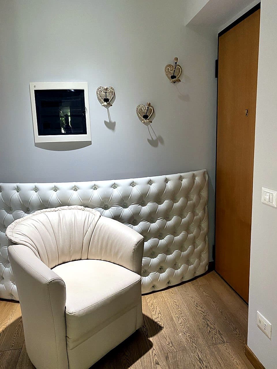 La piccola zona d'ingresso dell'appartamento · divanetto bianco capitonné, poltroncina tondeggiante, quadro elettrico a parete e la porta d'ingresso in legno chiaro a destra