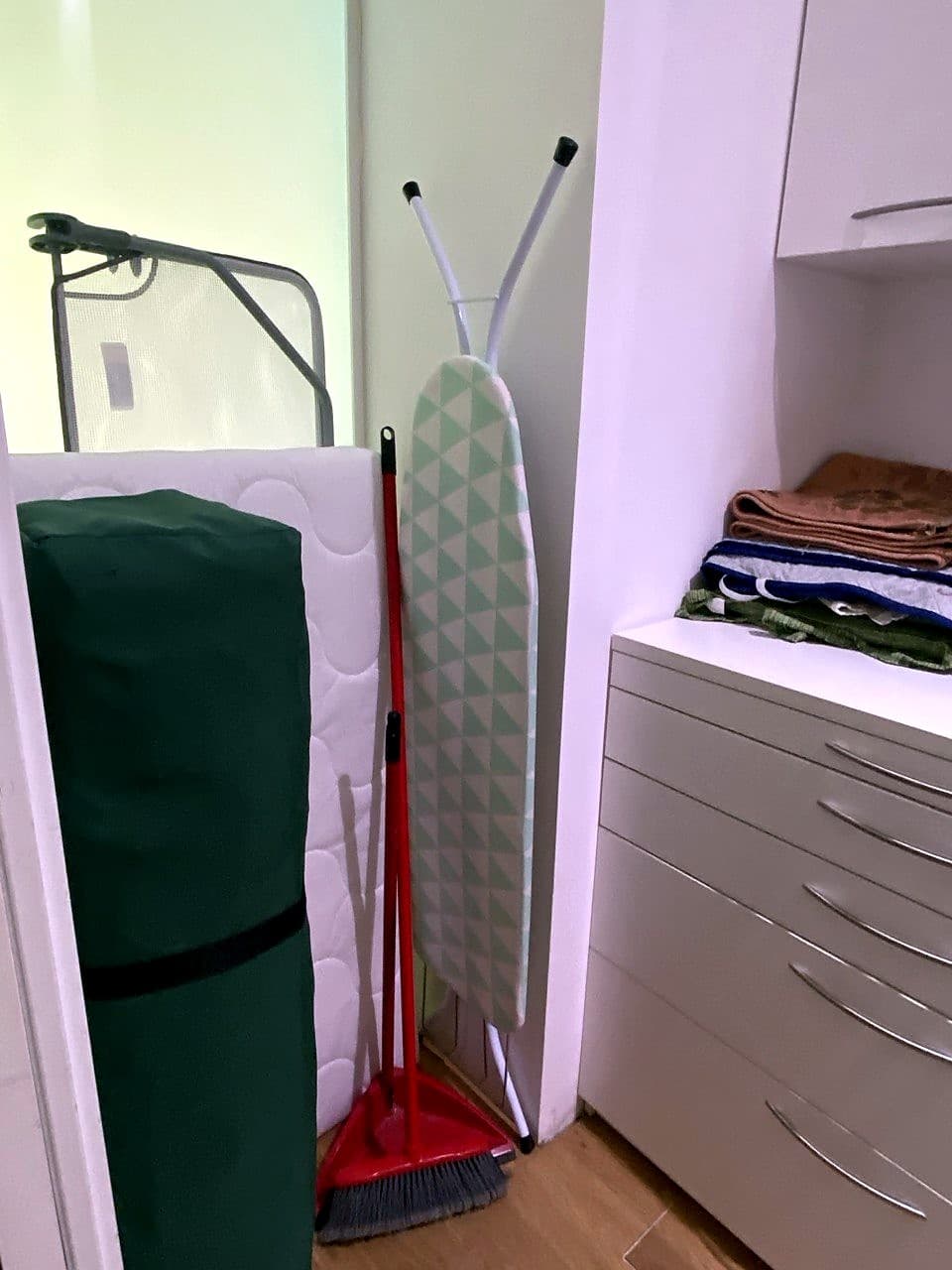 Angolo della lavanderia con asse da stiro, scopa, materassino da viaggio arrotolato e una colonna di cassetti con asciugamani e biancheria piegati
