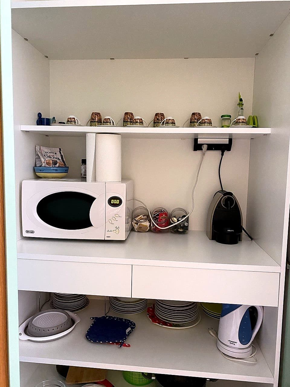 Lo sportello dell'armadio della cucina aperto · all'interno il microonde DeLonghi sopra, e sotto la dispensa con scorte di carta cucina, scatole di pasta, bicchieri di vetro, capsule del caffè