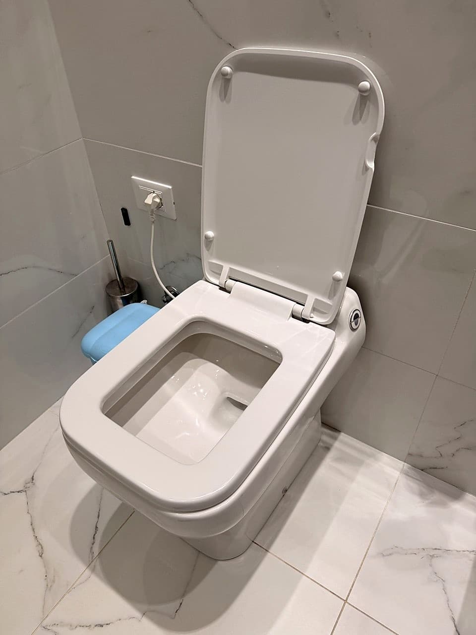 Il WC bianco moderno sospeso, con il coperchio sollevato · presa elettrica visibile dietro · pavimento in piastrelle effetto marmo